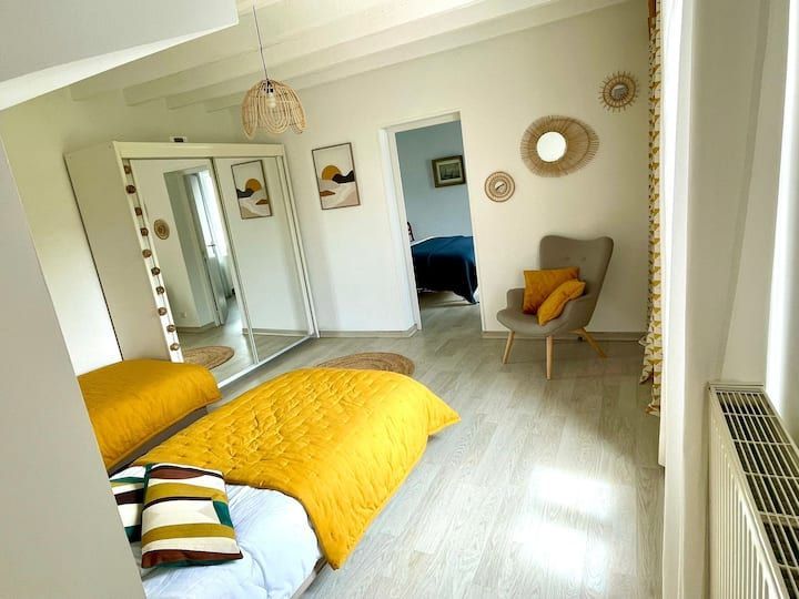 Chambre avec couvre-lit jaune, armoire blanche, murs aux tons neutres, miroirs en rotin et porte ouverte sur une autre pièce.