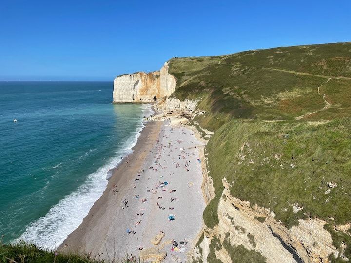 Scène de plage avec falaises blanches, collines verdoyantes, eau turquoise et personnes prenant un bain de soleil sur le sable.