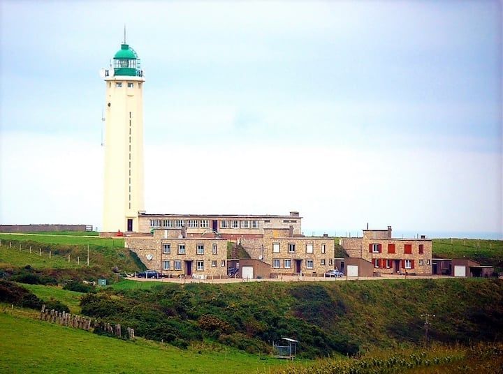 Grand phare blanc à sommet vert, à côté d'un groupe de bâtiments sur une falaise herbeuse.