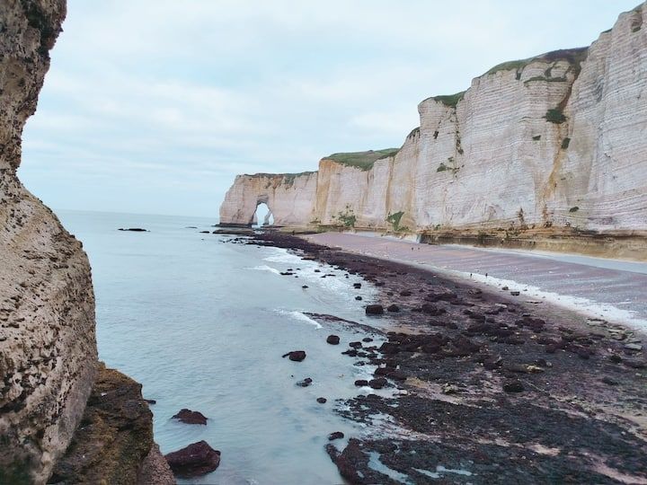 Falaises blanches surplombant une plage rocheuse et l'océan, avec une arche. Ciel nuageux.