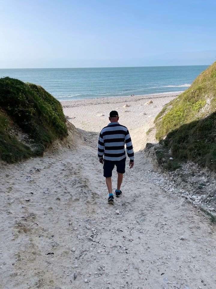 Un homme marche sur un chemin sablonneux en direction de l'océan, bordé de collines herbeuses sous un ciel bleu.