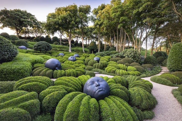 Des sculptures de visages émergeant de haies vertes taillées au cordeau dans un jardin avec un chemin et des arbres.