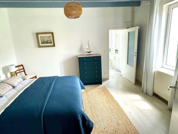 Chambre avec des touches de bleu : lit avec couverture bleue, commode verte, tapis, fenêtre, porte ouverte sur la salle de bain.