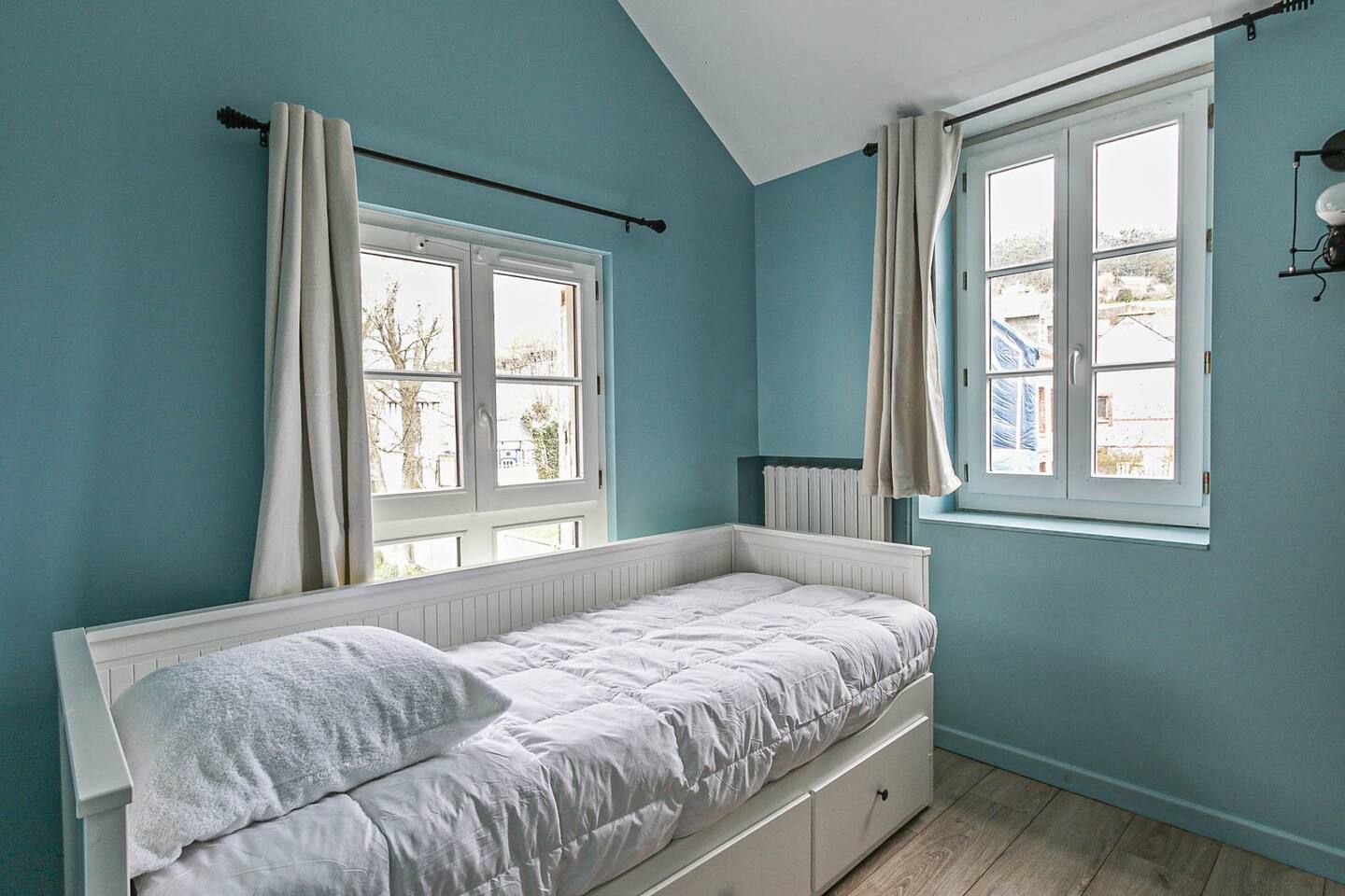 Une chambre aux murs bleus et un lit simple devant une fenêtre.