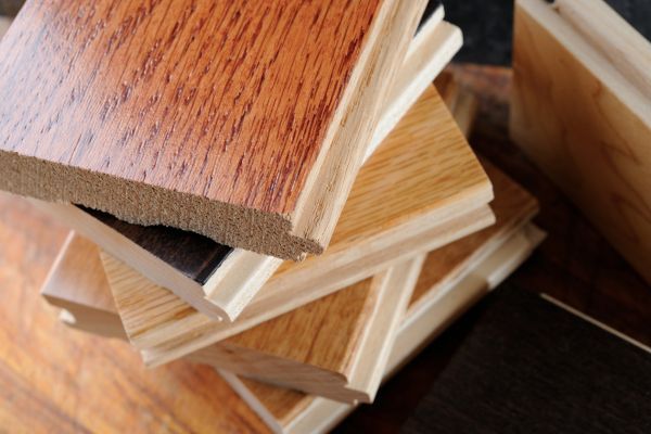 The image displays a stack of various hardwood flooring samples. Each sample showcases different wood species, colors, and grains, with textures ranging from smooth polished to textured matte finishes. The topmost sample has a reddish-brown hue with prominent wood grain, possibly cherry or mahogany. Below, there are lighter samples, likely oak or maple, with natural and honey-stained finishes. The samples are piled haphazardly, indicating a selection process, perhaps for flooring choices. They are set against a dark background that contrasts with the wood, highlighting the different wood tones and finishes.