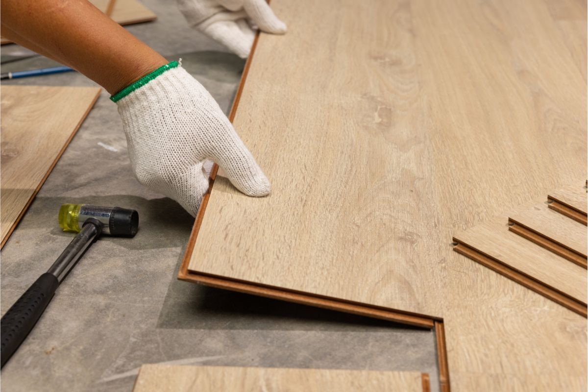 In Athens, GA, a worker’s hand, protected by a white fabric glove, is carefully placing a plank of light oak laminate flooring. The plank edges are interlocking with an existing series of laid planks on a grey underlayment. To the left, a black rubber mallet, indicating recent use, rests on the concrete floor. Scattered around are more laminate pieces awaiting installation. The scene highlights the precision required in flooring work, with the focus on the alignment of the plank being secured into place.