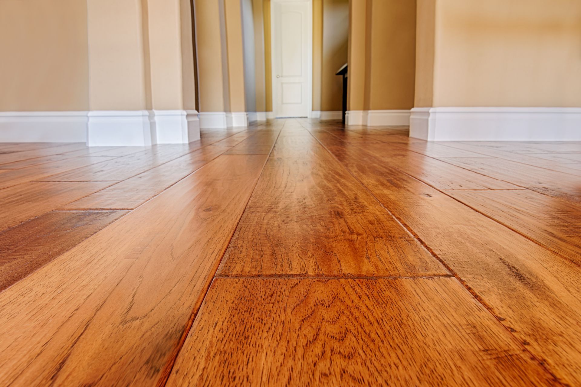 The image displays the detailed craftsmanship of hardwood flooring in an Athens, GA, home. The warm, amber tones of the polished wood planks stretch across the room, leading to a white door. The wood grain, knots, and seams add character and texture, complementing the white baseboard and soft beige walls. The perspective emphasizes the expanse of the floor, showcasing its glossy finish and the meticulous installation that enhances the room's welcoming ambiance.