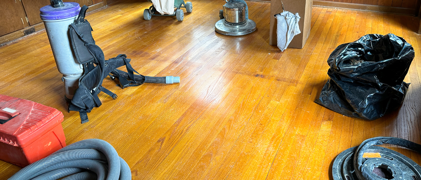 A photo of hardwood refinishing tools on a hardwood floor in a house. The floor is visibly worn down and desperately needs a refinishing job.