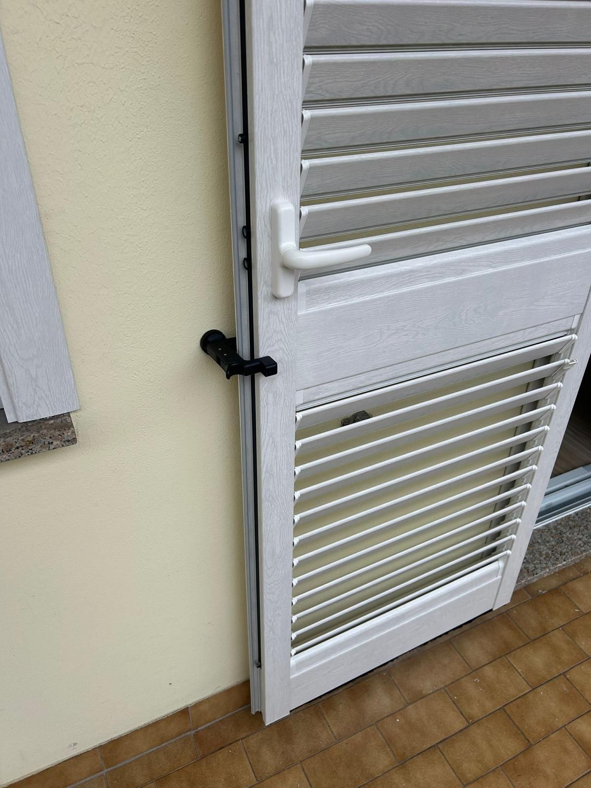 Persiana bianca con persiana, parzialmente aperta, con maniglia e chiavistello, montata su una parete color crema.