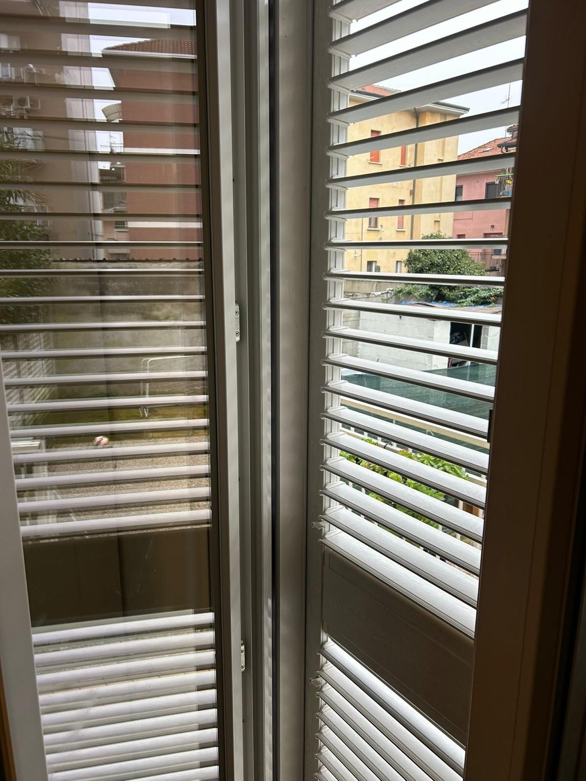 Vista interna delle persiane bianche sopra la finestra che si affaccia su una strada con edifici.