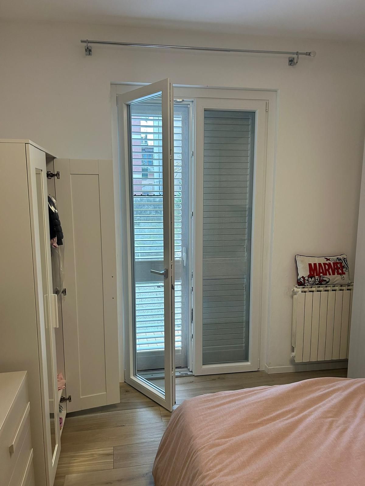 Camera da letto con porta bianca aperta che conduce al balcone, armadio bianco sulla sinistra, letto rosa e termosifone.