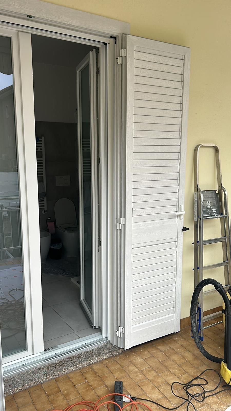Porta bianca e persiana su un portico, parzialmente aperte, che rivelano un bagno. Accanto alla persiana ci sono una scala e un aspirapolvere.
