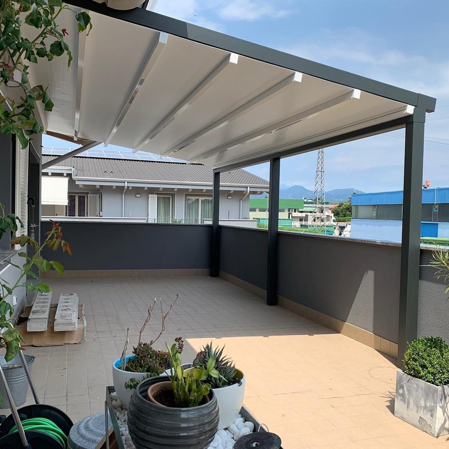 Terrazza con tettoia bianca retrattile, struttura grigio scuro, piante in vaso e vista sulla città.