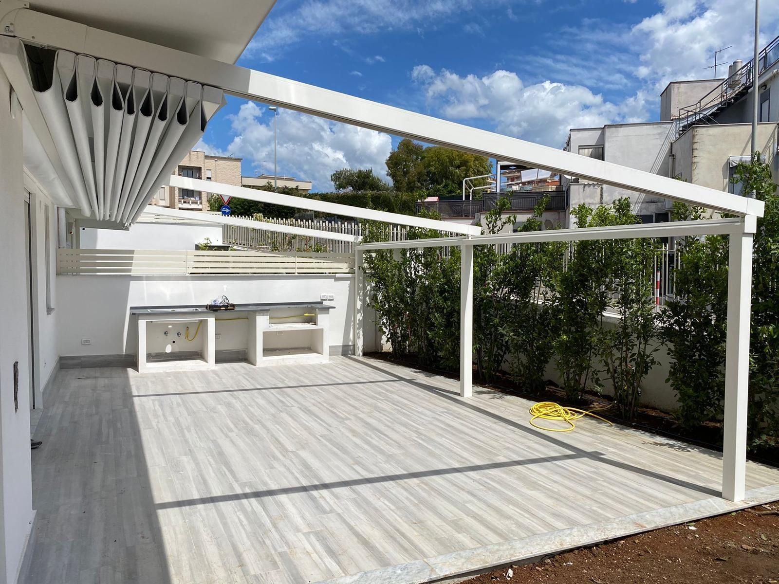 Pergola bianca sopra un patio con cucina esterna integrata. Cielo azzurro sullo sfondo.