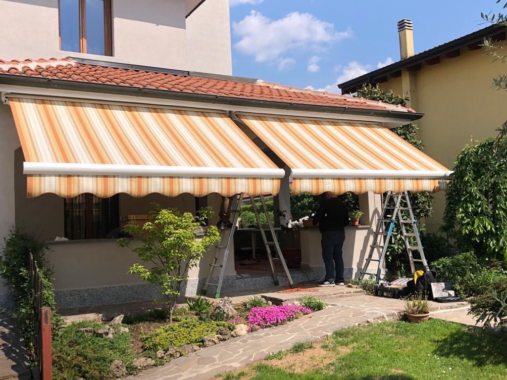 Due tende a strisce sul portico di una casa, una persona su una scala le sta installando, circondate da piante.