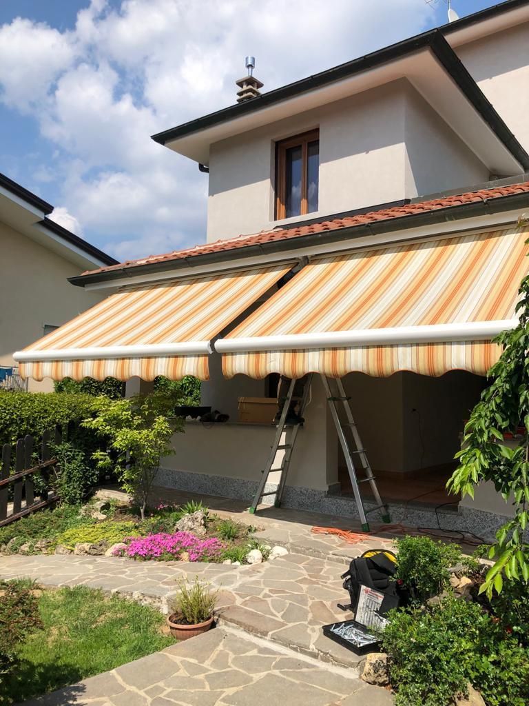 Due tende a strisce su una casa beige, con un giardino antistante. Una scala è appoggiata al muro.