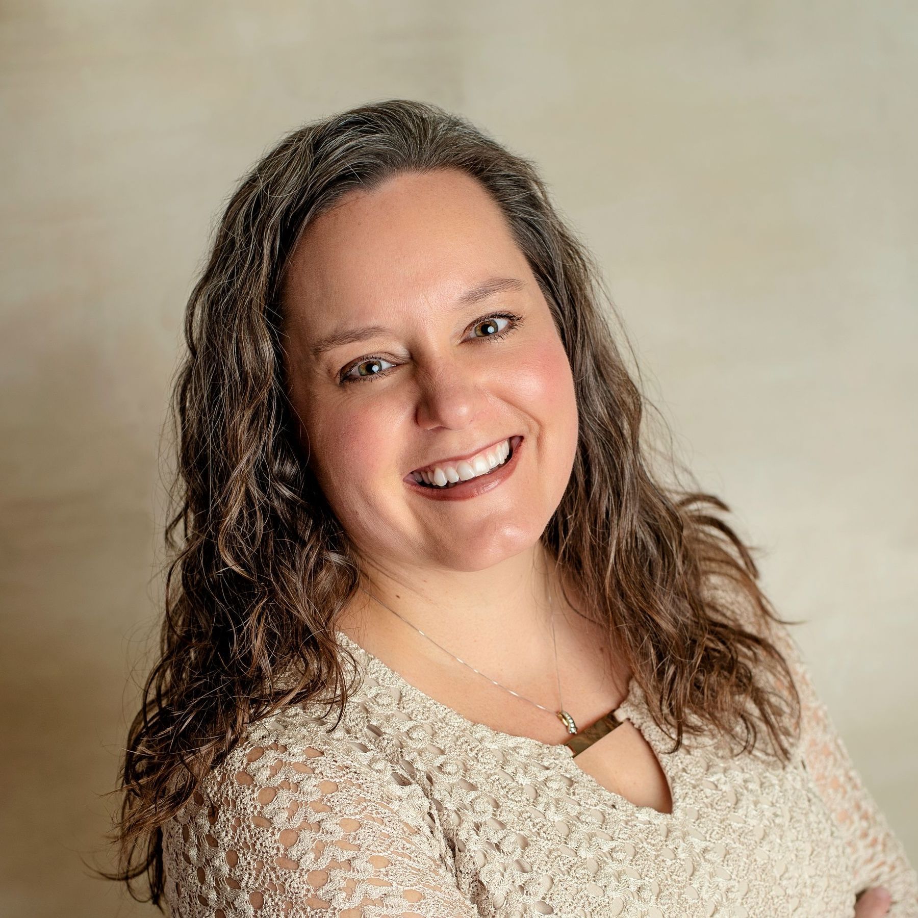 Woman with brown hair smiles, wearing a beige patterned top, posing in front of a neutral background.