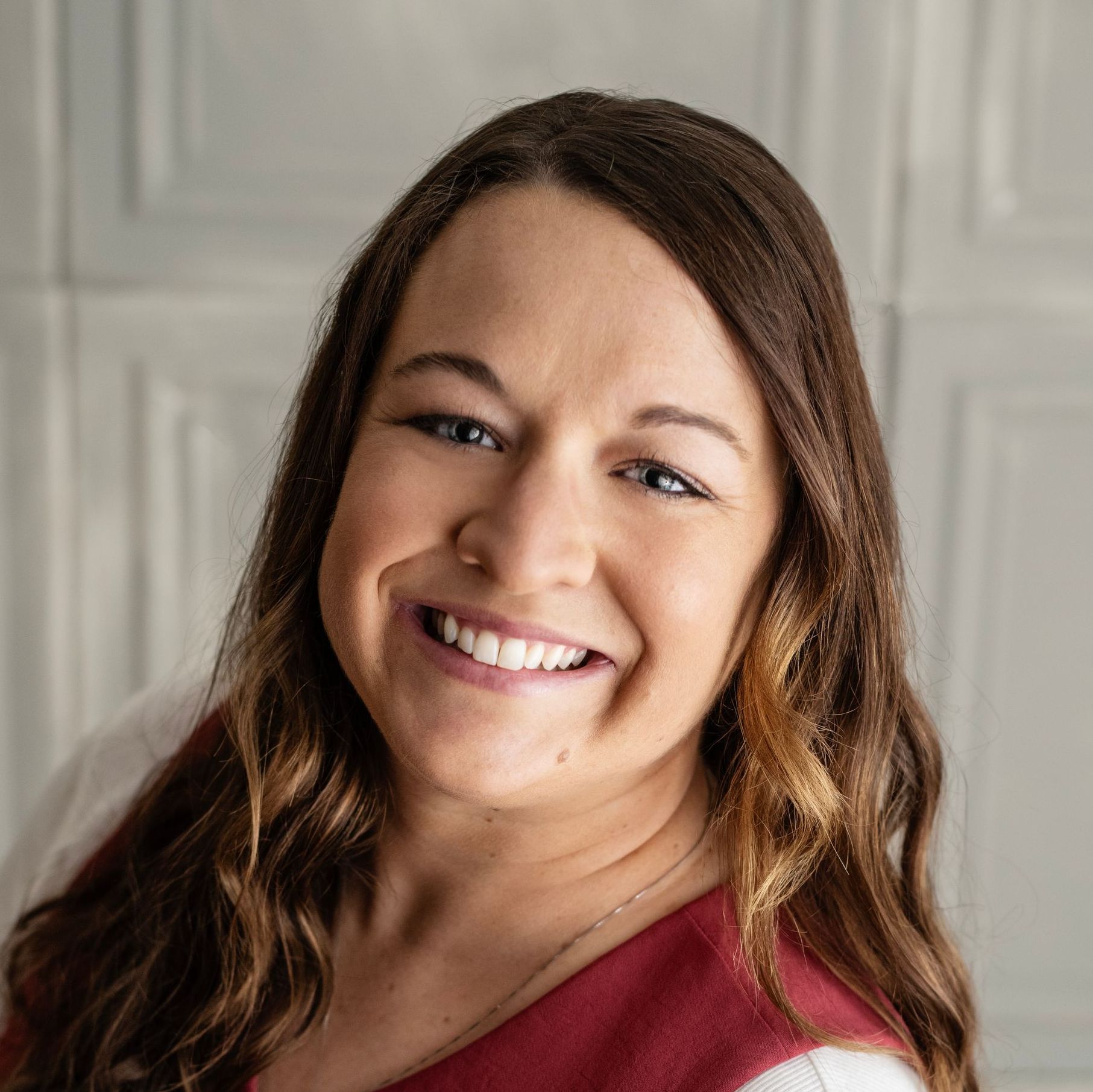 Woman with long brown hair smiles at the camera, wearing a red top against a white paneled background.
