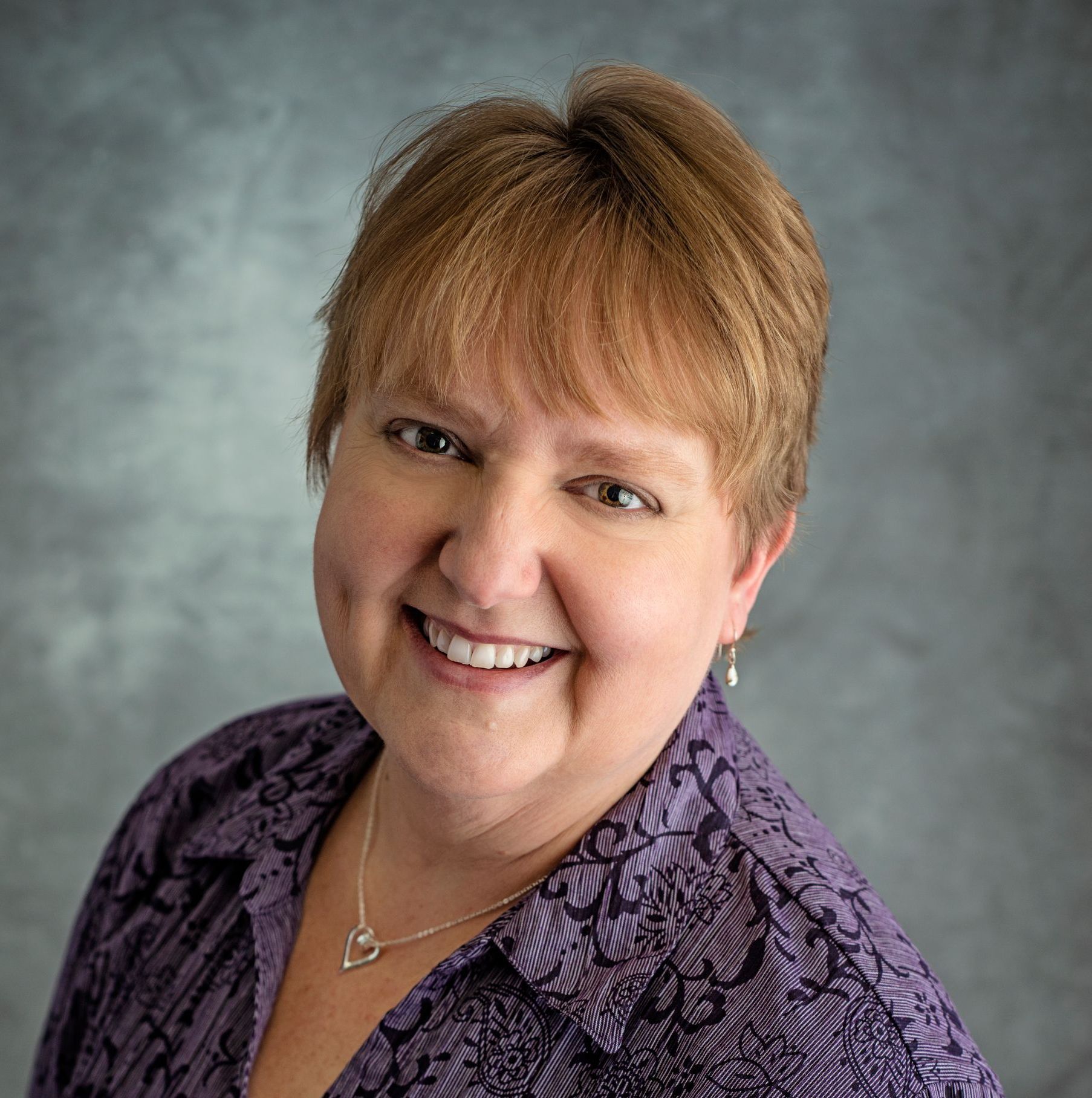 Woman with short, light brown hair smiles, wearing a purple floral button-down shirt and silver necklace.
