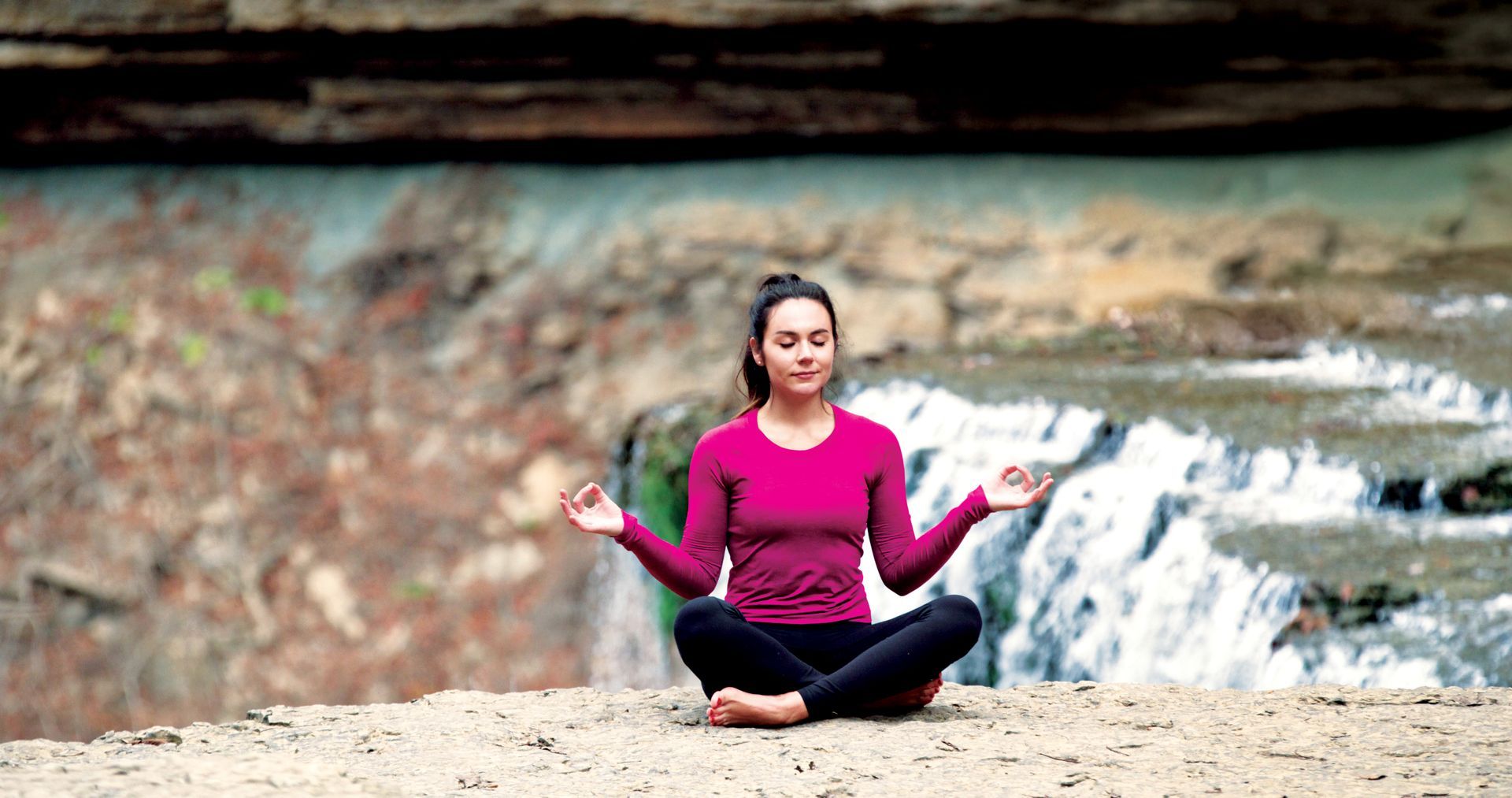 Woman in lotus pose, eyes closed, meditating near a waterfall. Wearing pink top and black pants.
