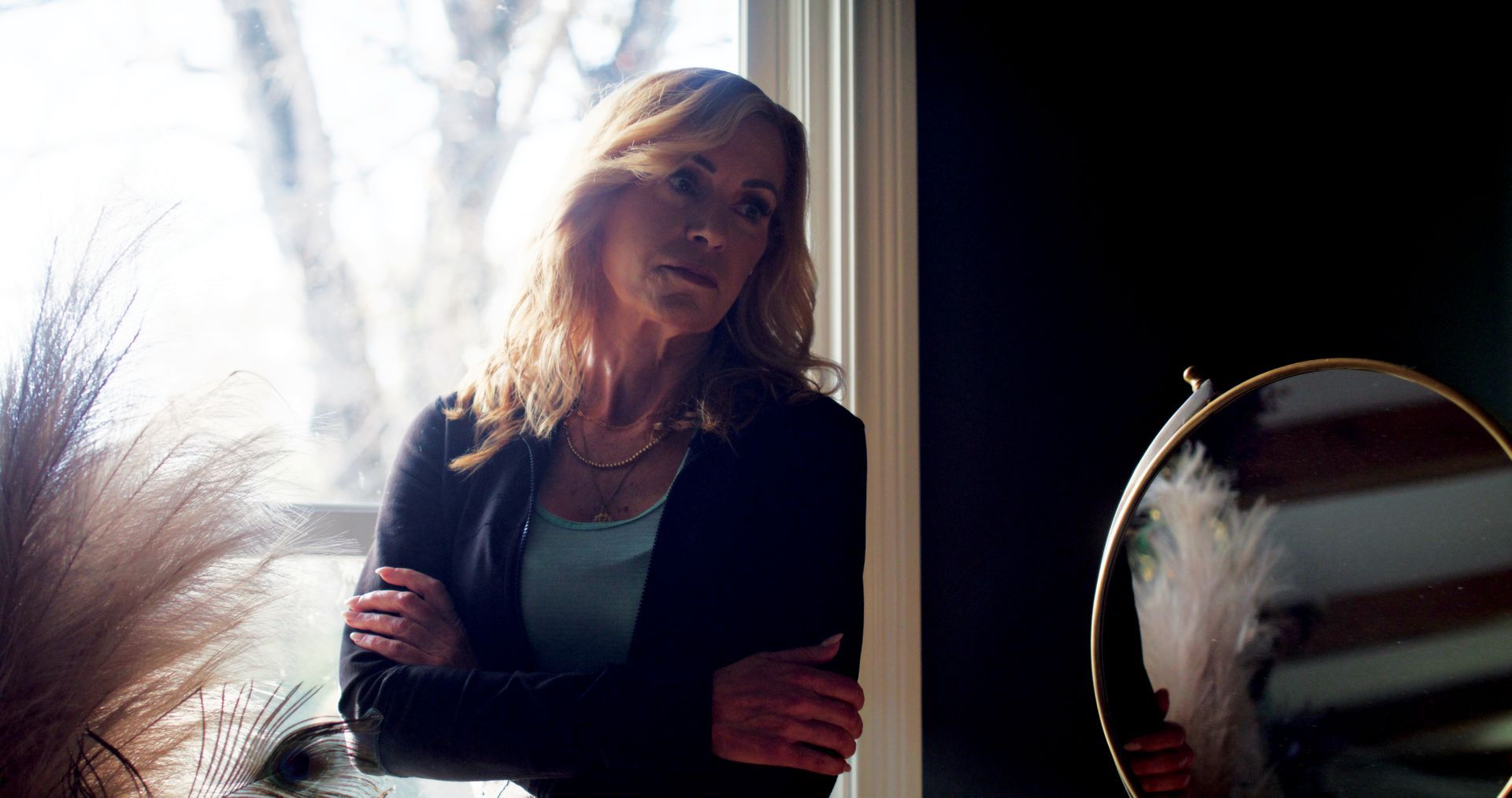 Woman in a blazer looks pensively out a window, arms crossed. A small mirror is in the foreground.