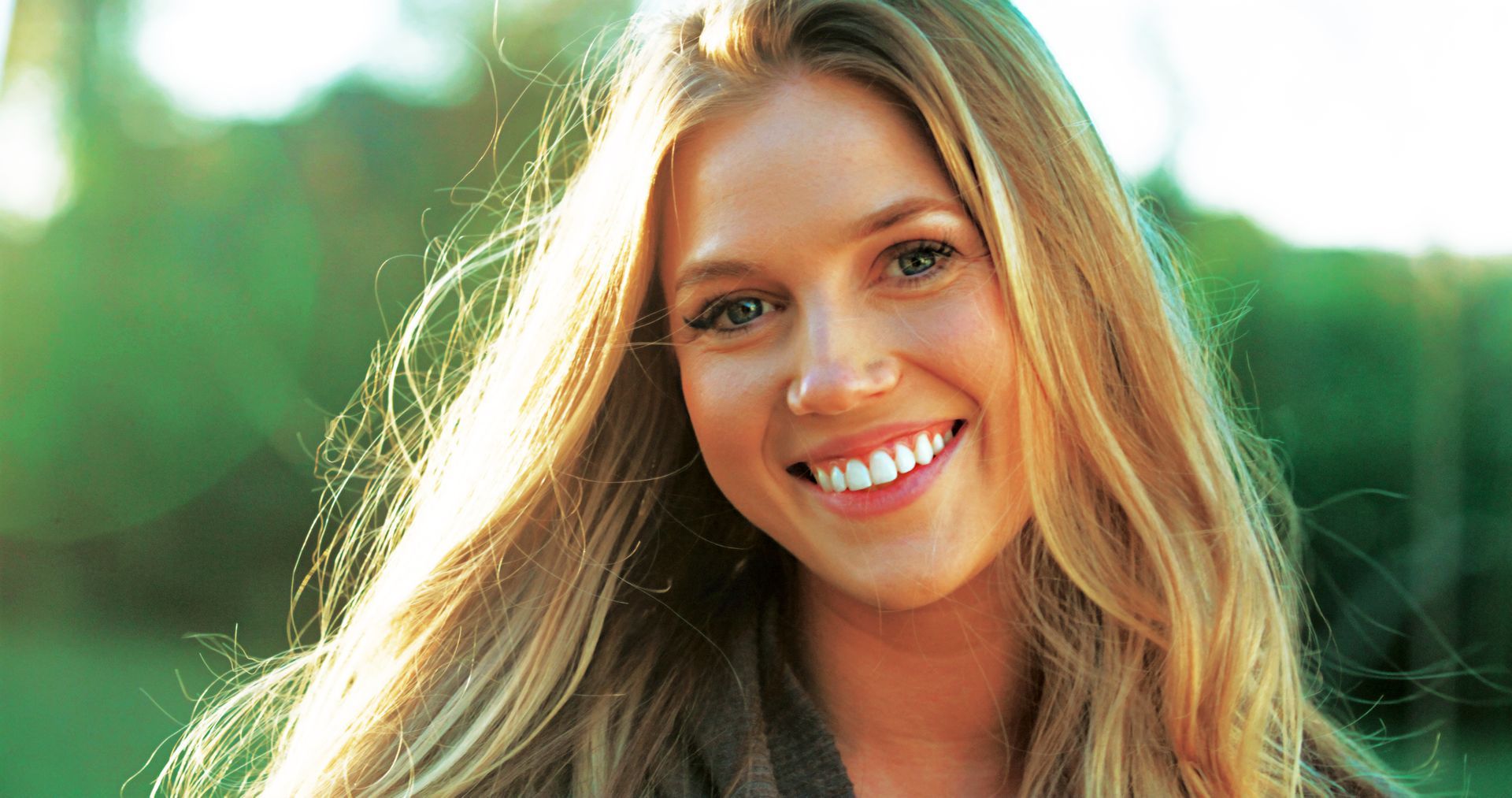 Blonde woman smiling, lit by sunlight, outdoors.