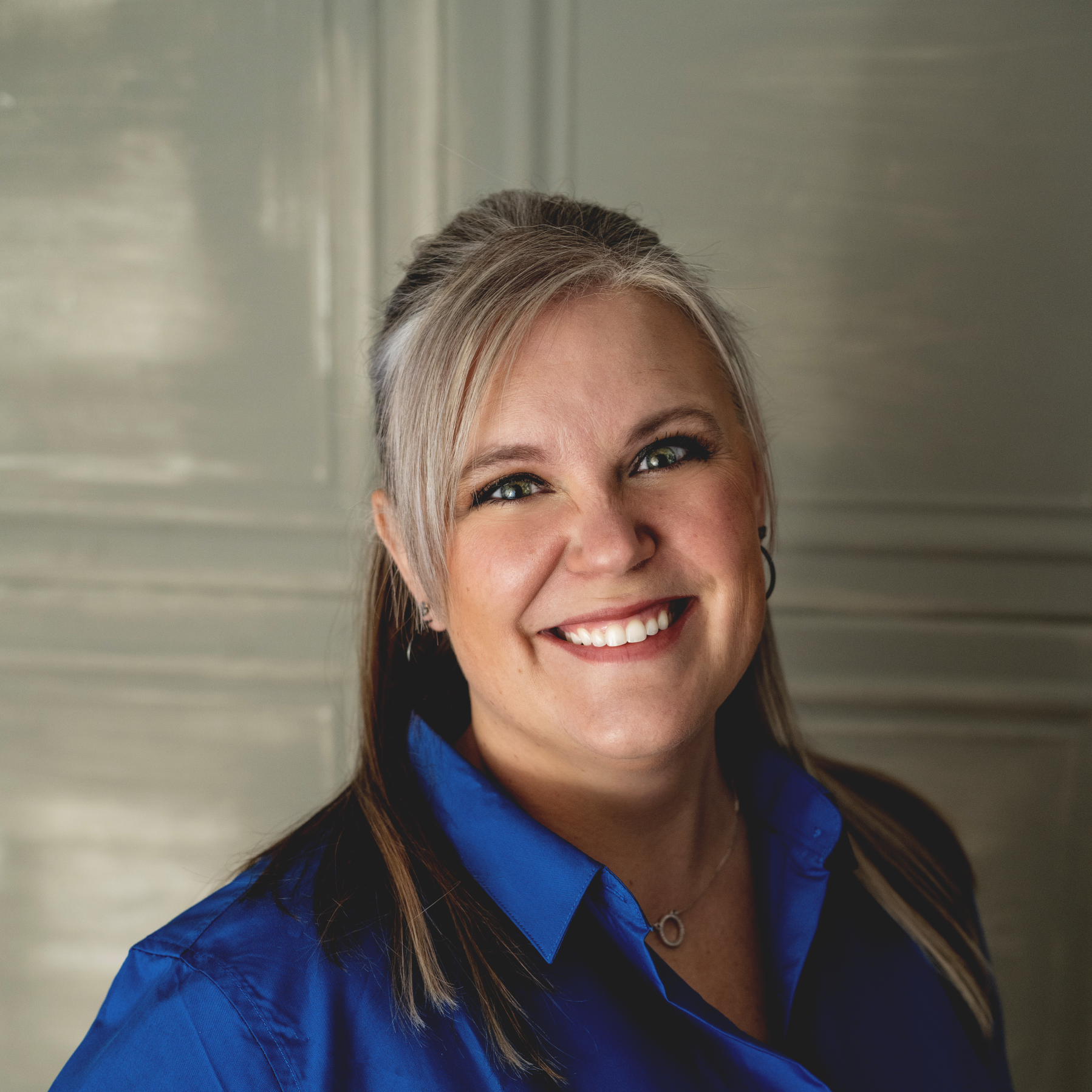 Woman with long brown hair smiles at the camera, wearing a red top against a white paneled background.