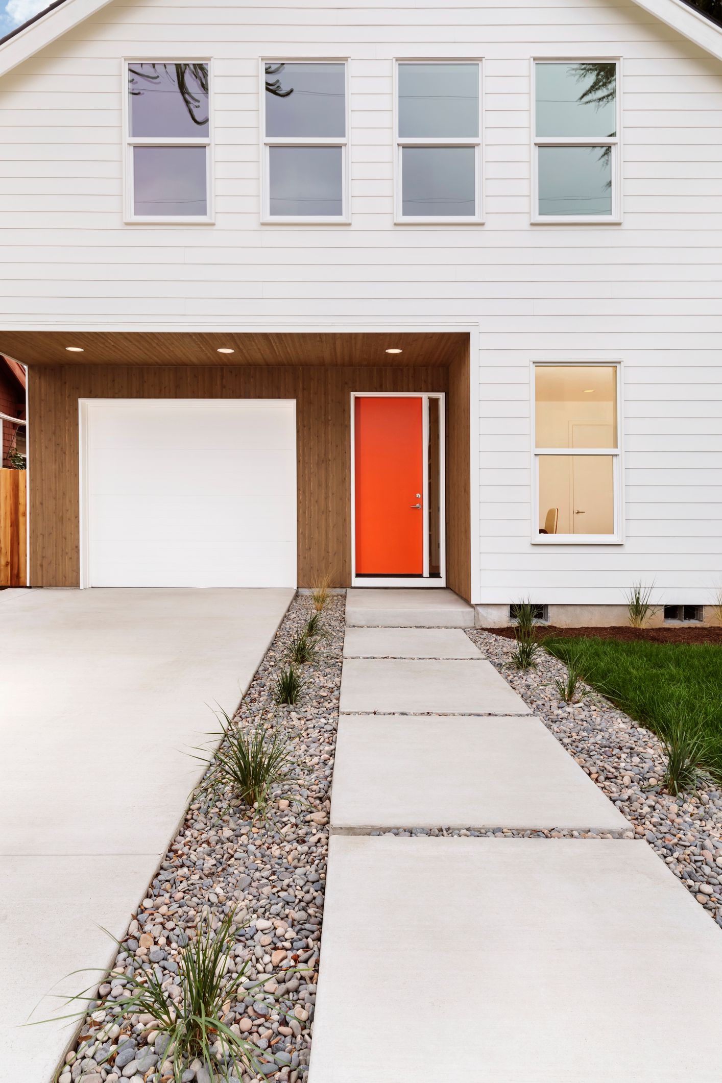 White house with orange door, garage, and a concrete pathway with rock gardens.