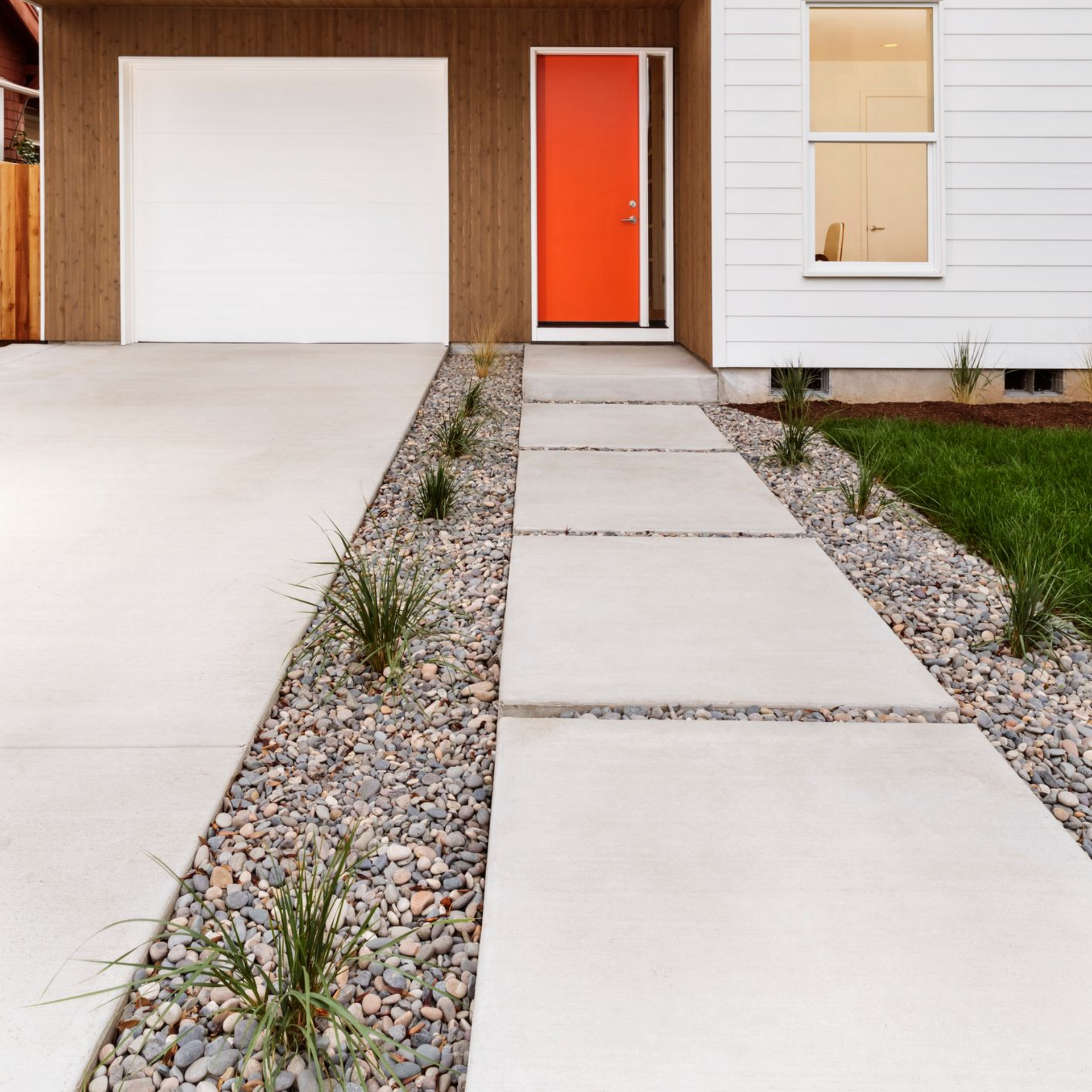 White house with orange door, garage, and a concrete pathway with rock gardens.