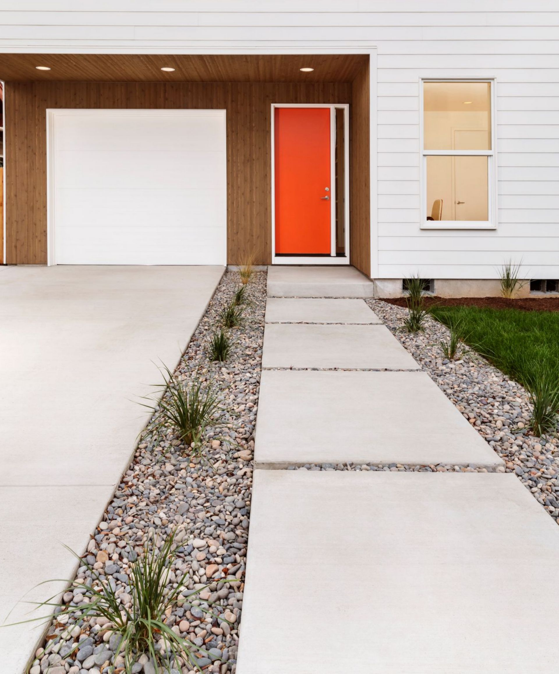 White house with orange door, garage, and a concrete pathway with rock gardens.