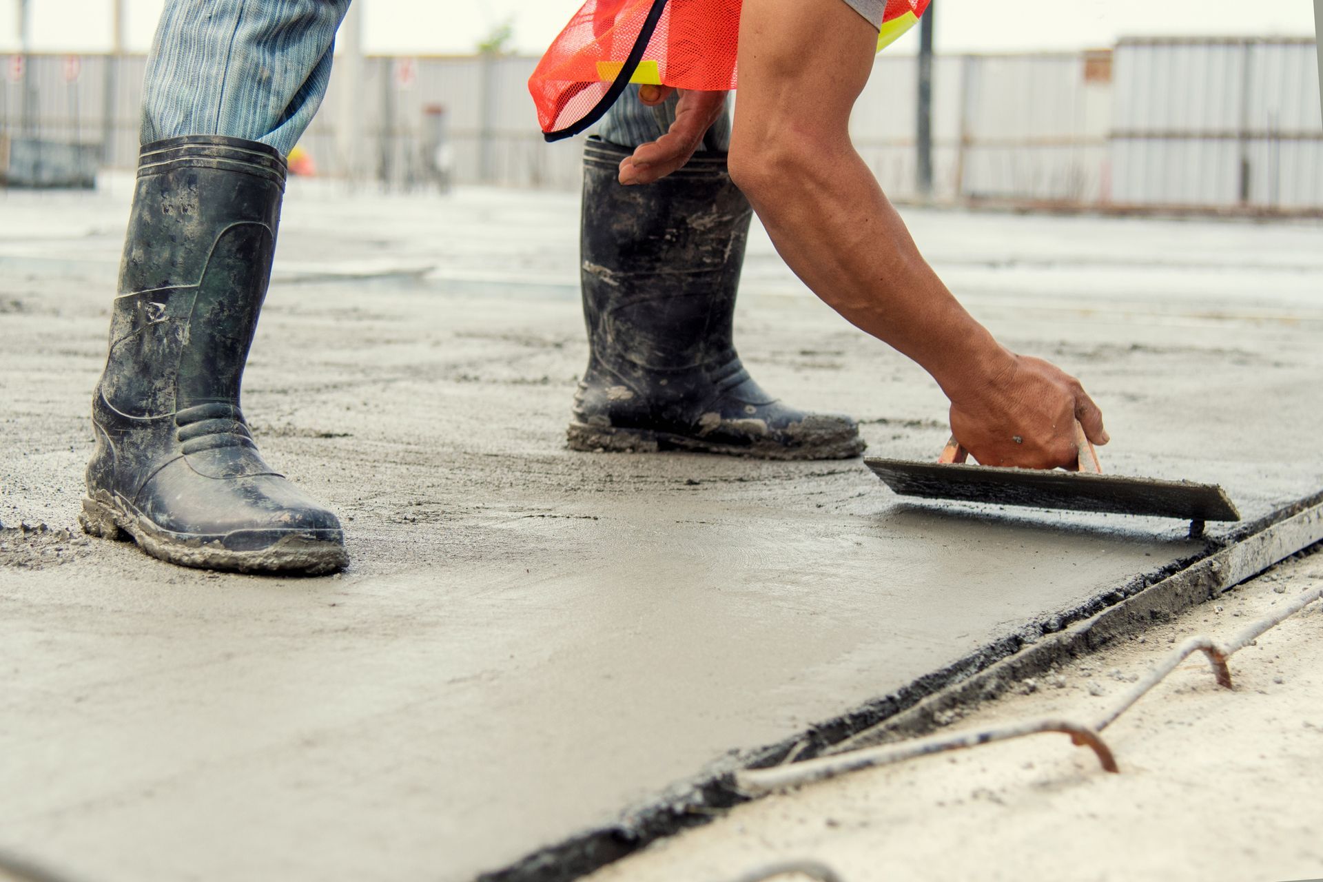 Construction worker using a trowel to smooth wet concrete. Boots, orange vest, outdoor setting.