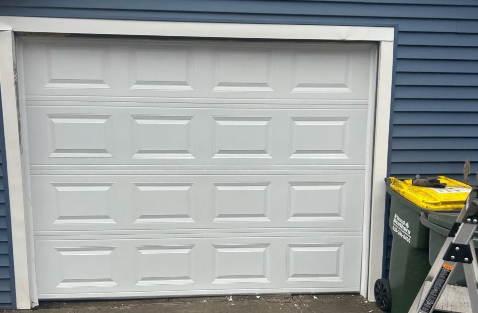 A white garage door is open next to a green trash can and a ladder.