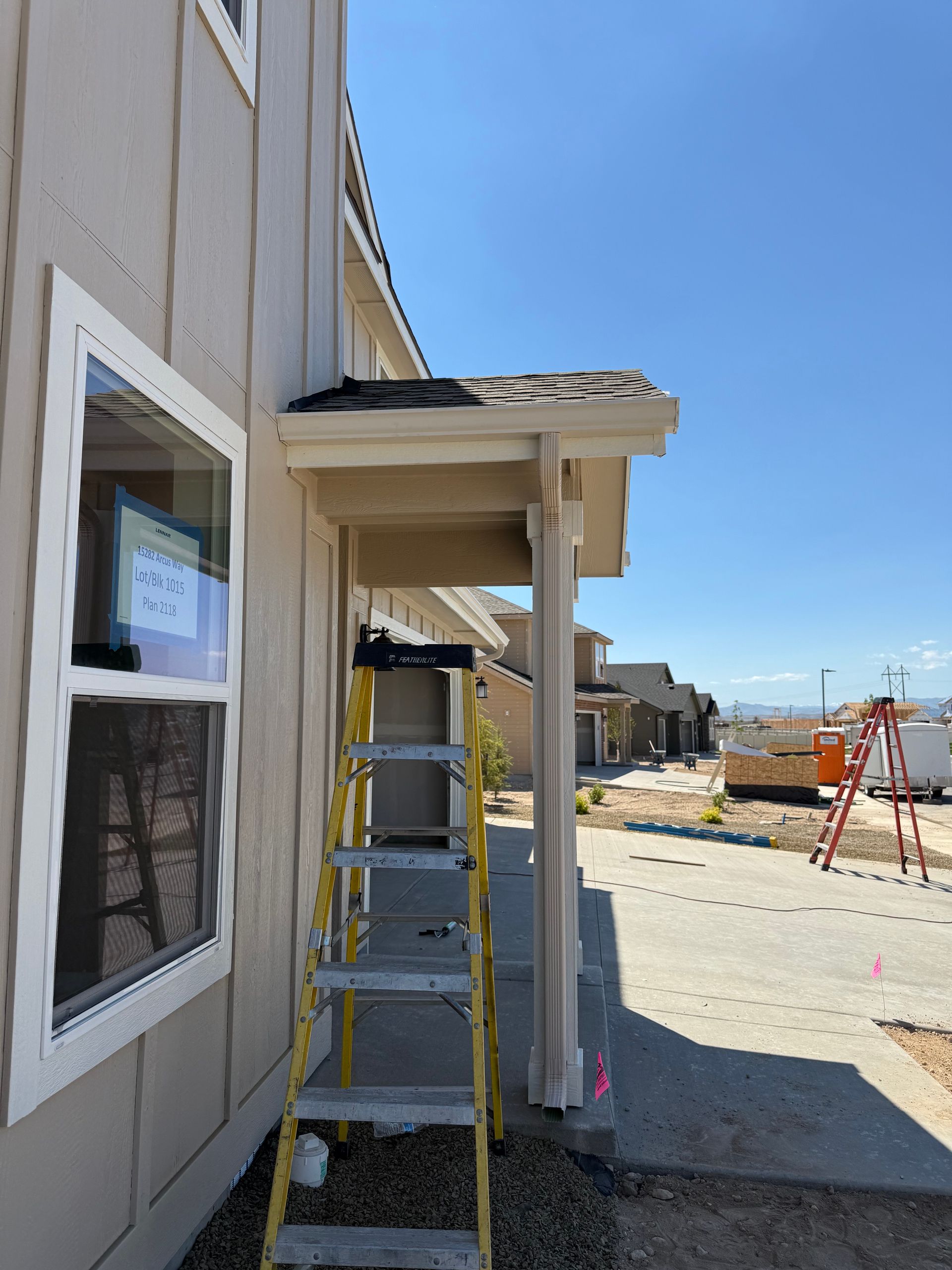 Ladder leans against a beige house with white window and a covered porch. Construction site setting.