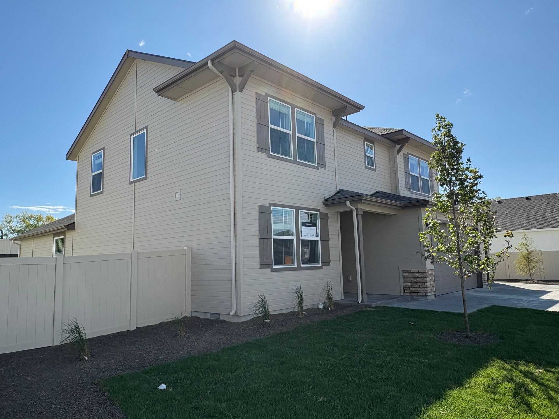 Two-story beige house with brown shutters and a small front yard, under a bright blue sky.