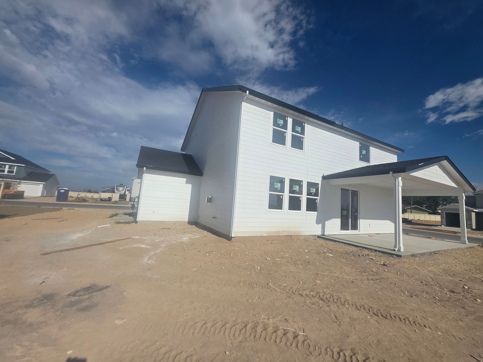 New white two-story house under construction with a black roof and attached patio on a dirt lot, blue sky.