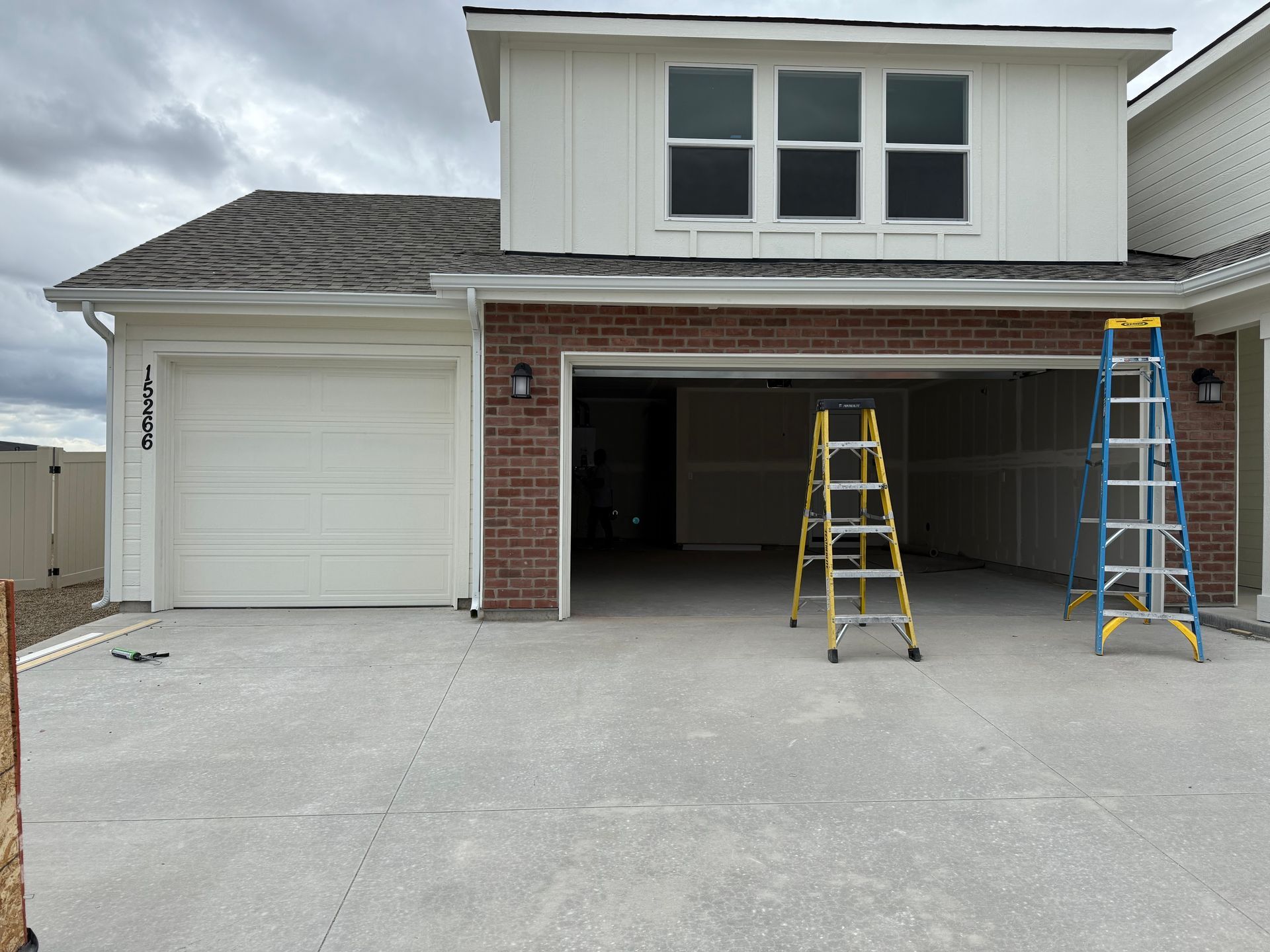 Two-car garage with open bays. Red brick, white siding, and two ladders present. Cloudy sky overhead.