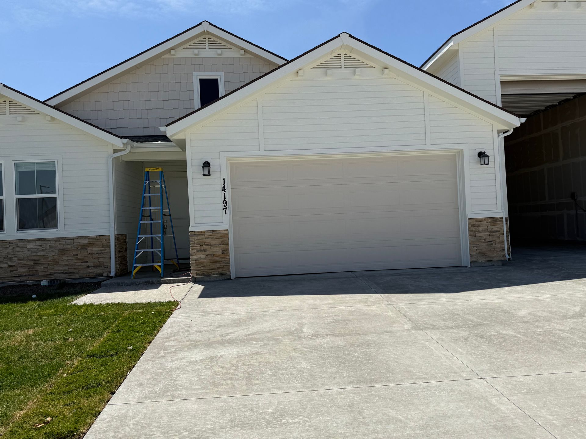 White house with a tan garage door, brown stone accents, and a ladder by the front door on a concrete driveway.