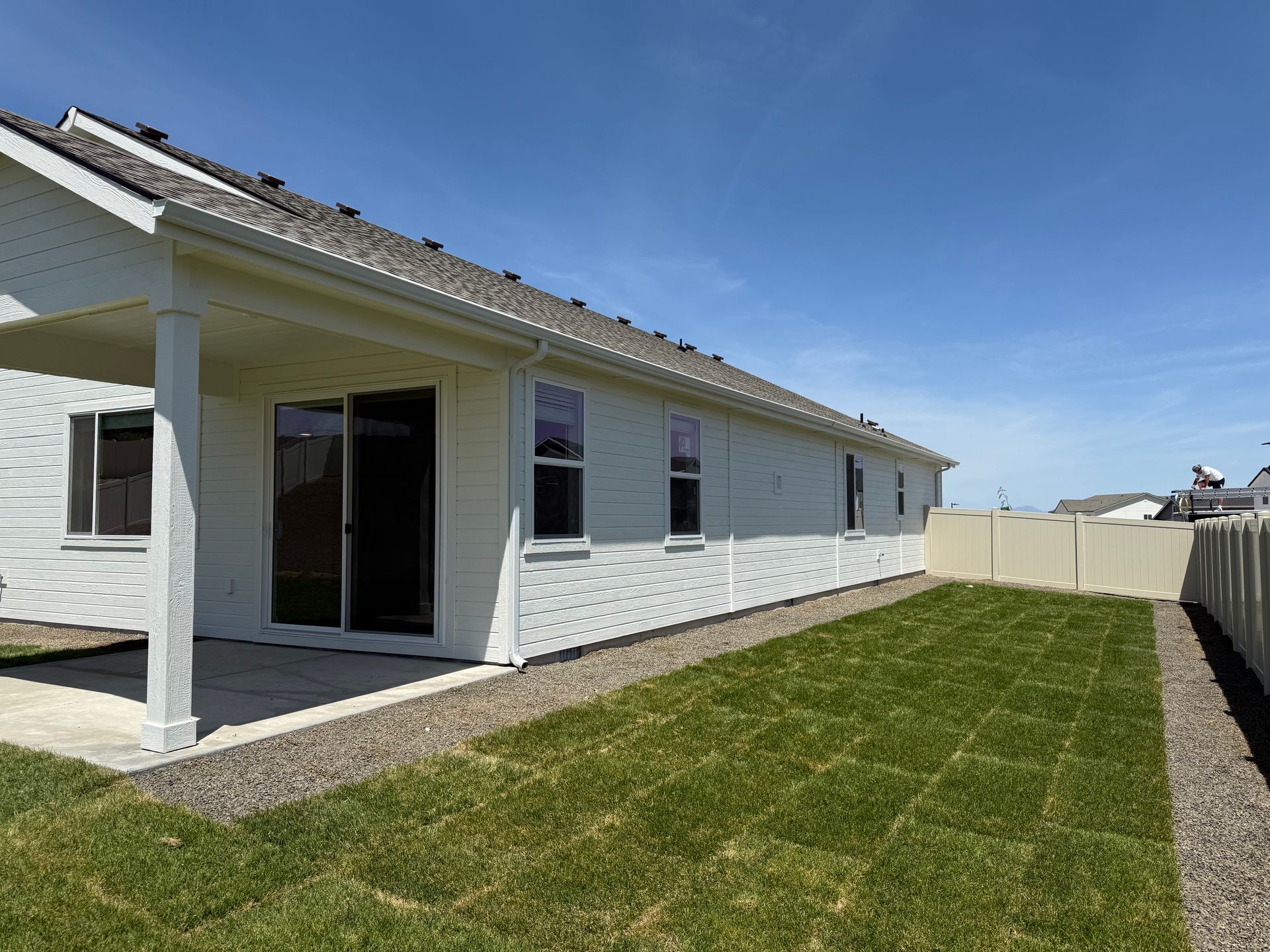 Backyard of a white house with a covered patio, grass, and a beige fence under a blue sky.