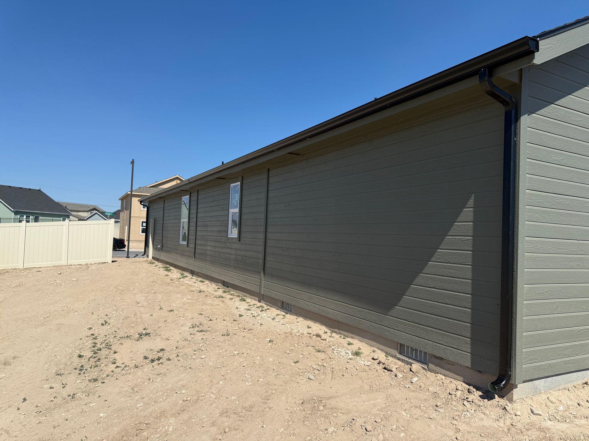 Exterior view of a house with gray siding, black gutters, and windows. Brown dirt yard. Blue sky.