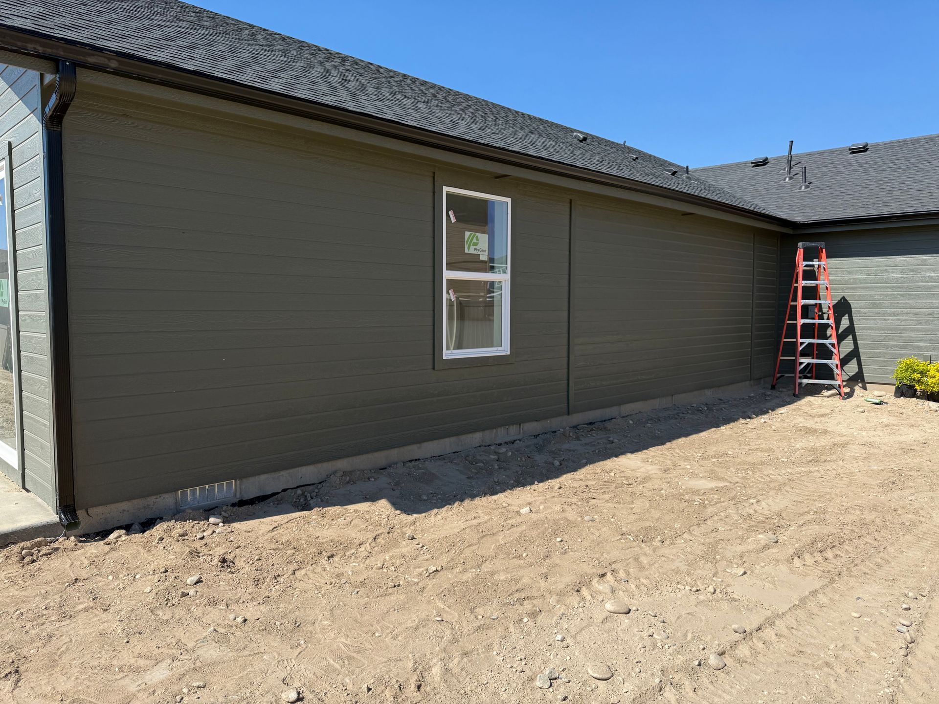 Exterior wall of a house with green siding, a window, and a ladder on a dirt lot.