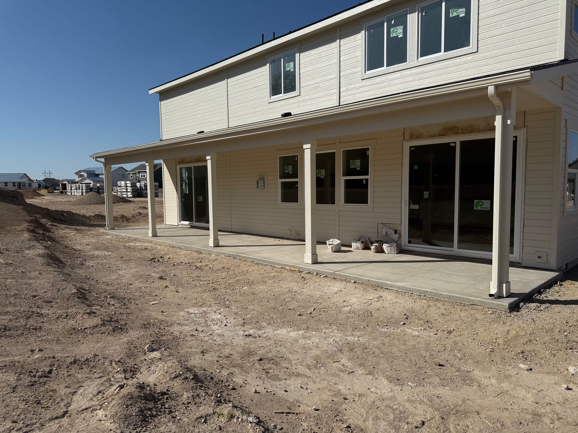 Back of a two-story beige house under construction with a long covered patio and bare dirt.