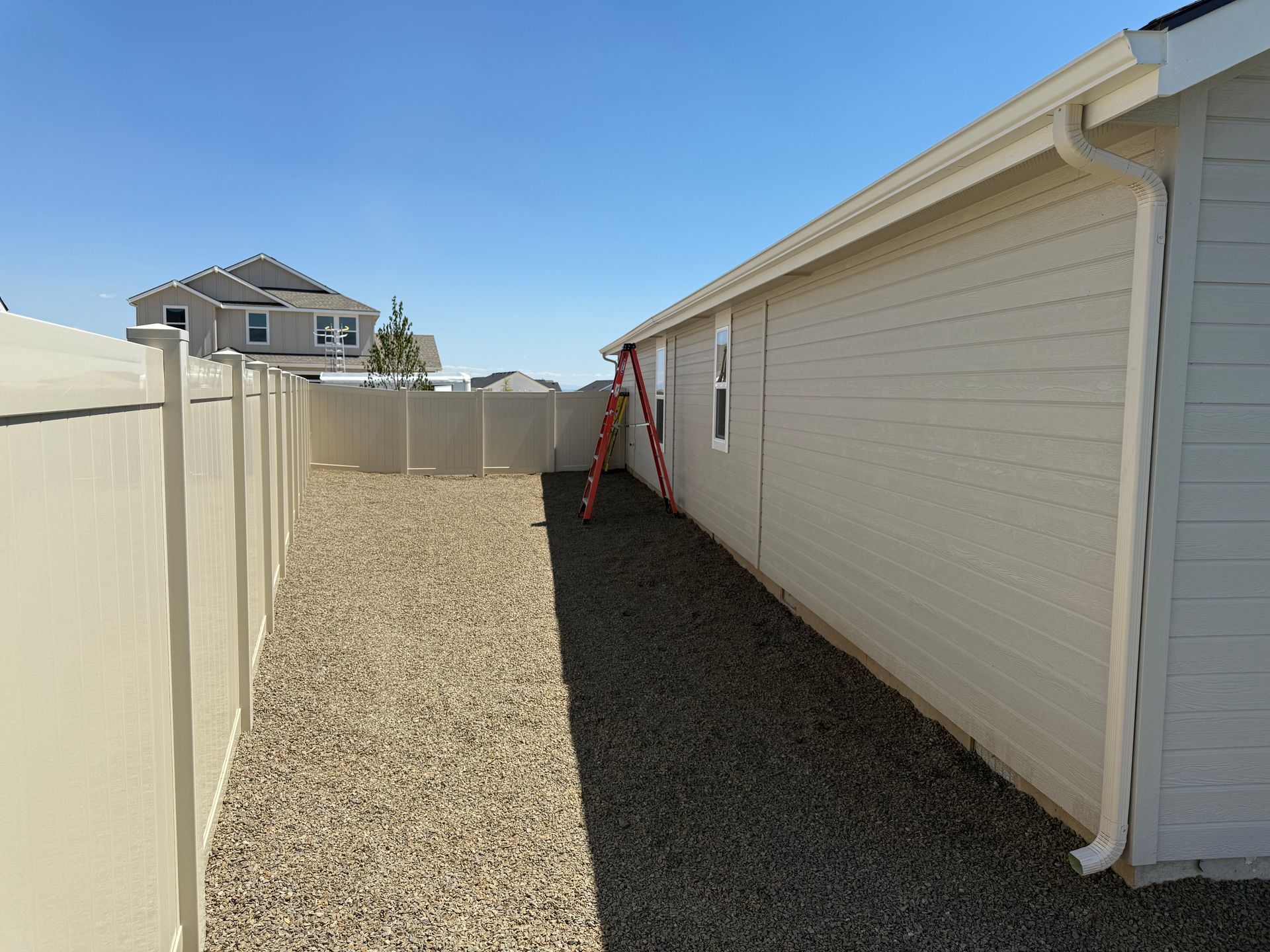 Ladder leans against a beige building. Beige fence and gravel path. Sunny sky.