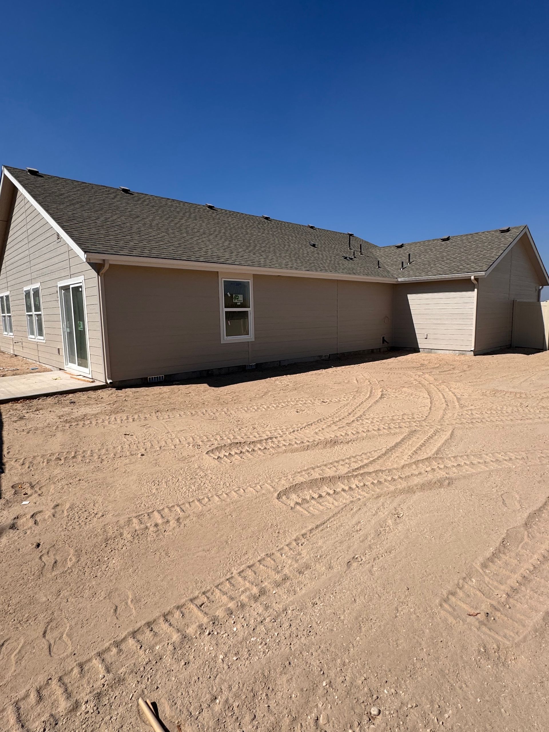 Tan house under construction with brown roof against a clear, blue sky. Sandy ground and tire tracks in front.