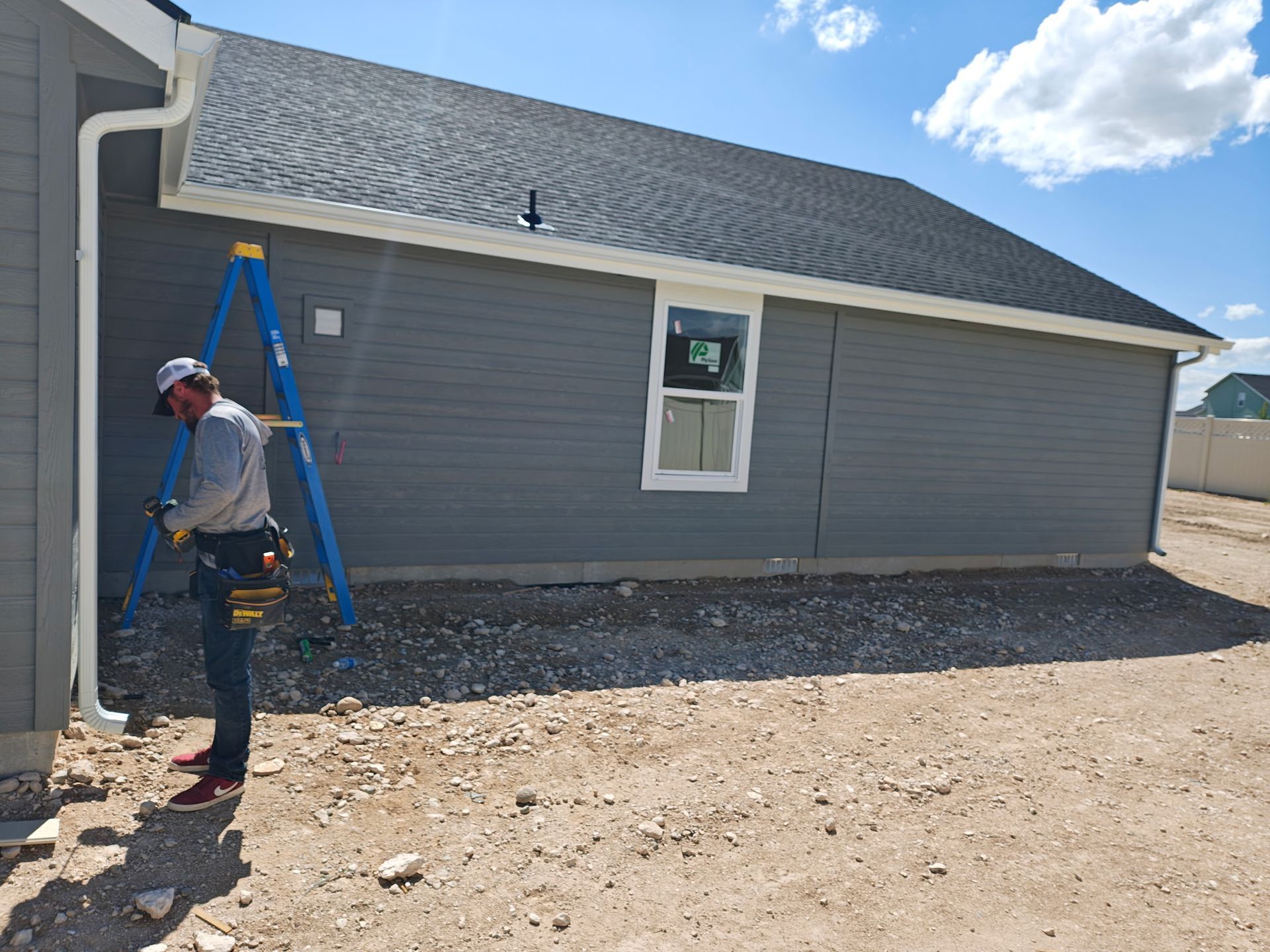 A worker installs a gutter on a gray-sided house. A blue ladder, gravel ground, and sunny sky are present.