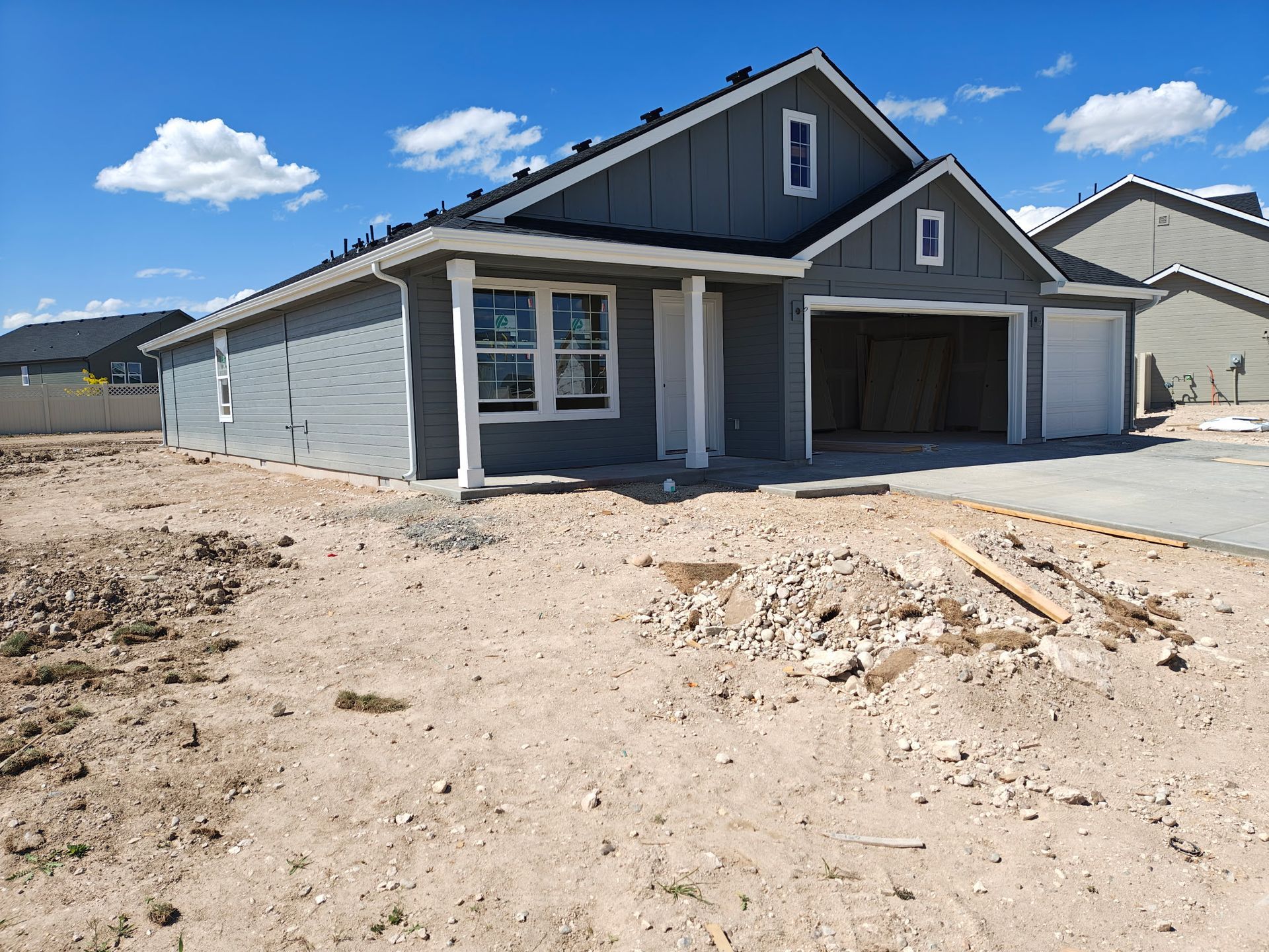 New gray house under construction, with open garage, on a dirt lot, blue sky.