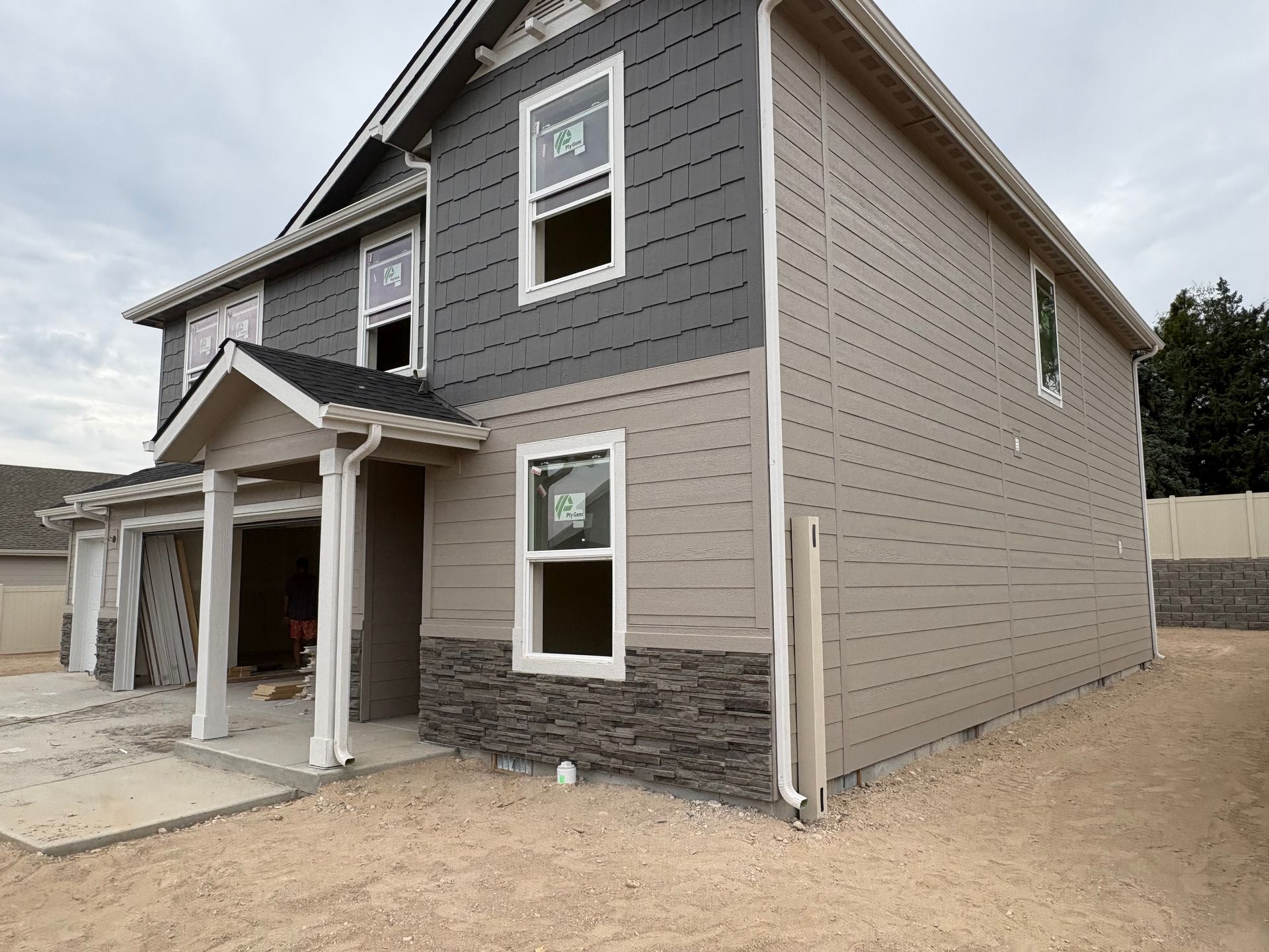 Two-story house under construction with gray and beige siding, stone accents, and unfinished landscaping.