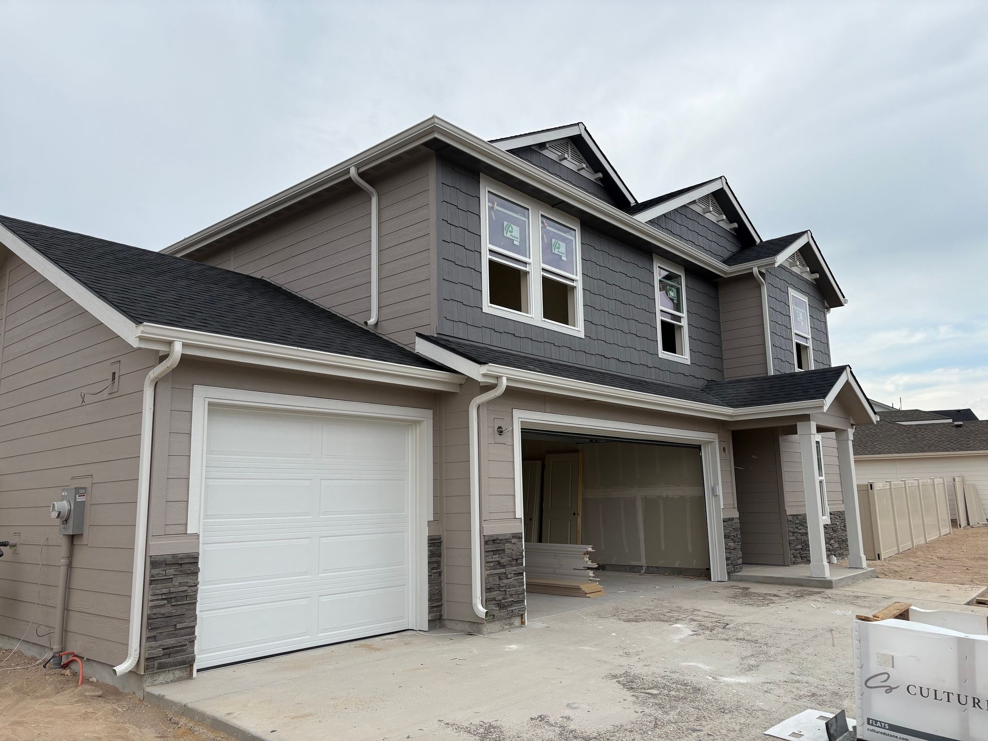 New construction two-story house with gray and tan siding, white garage doors, and a cloudy sky.