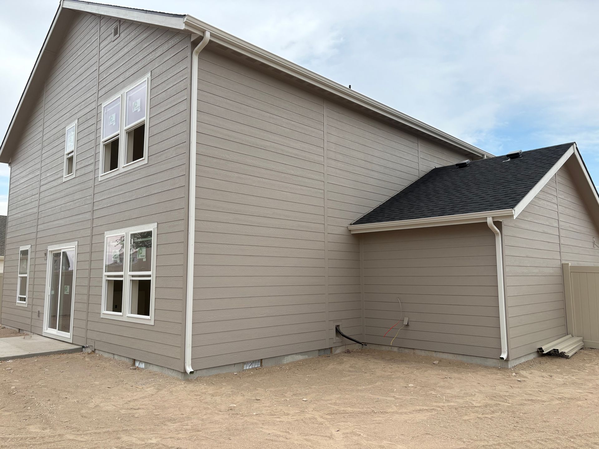 Beige two-story house with white trim, windows, and attached garage with black roof on a gravel lot.