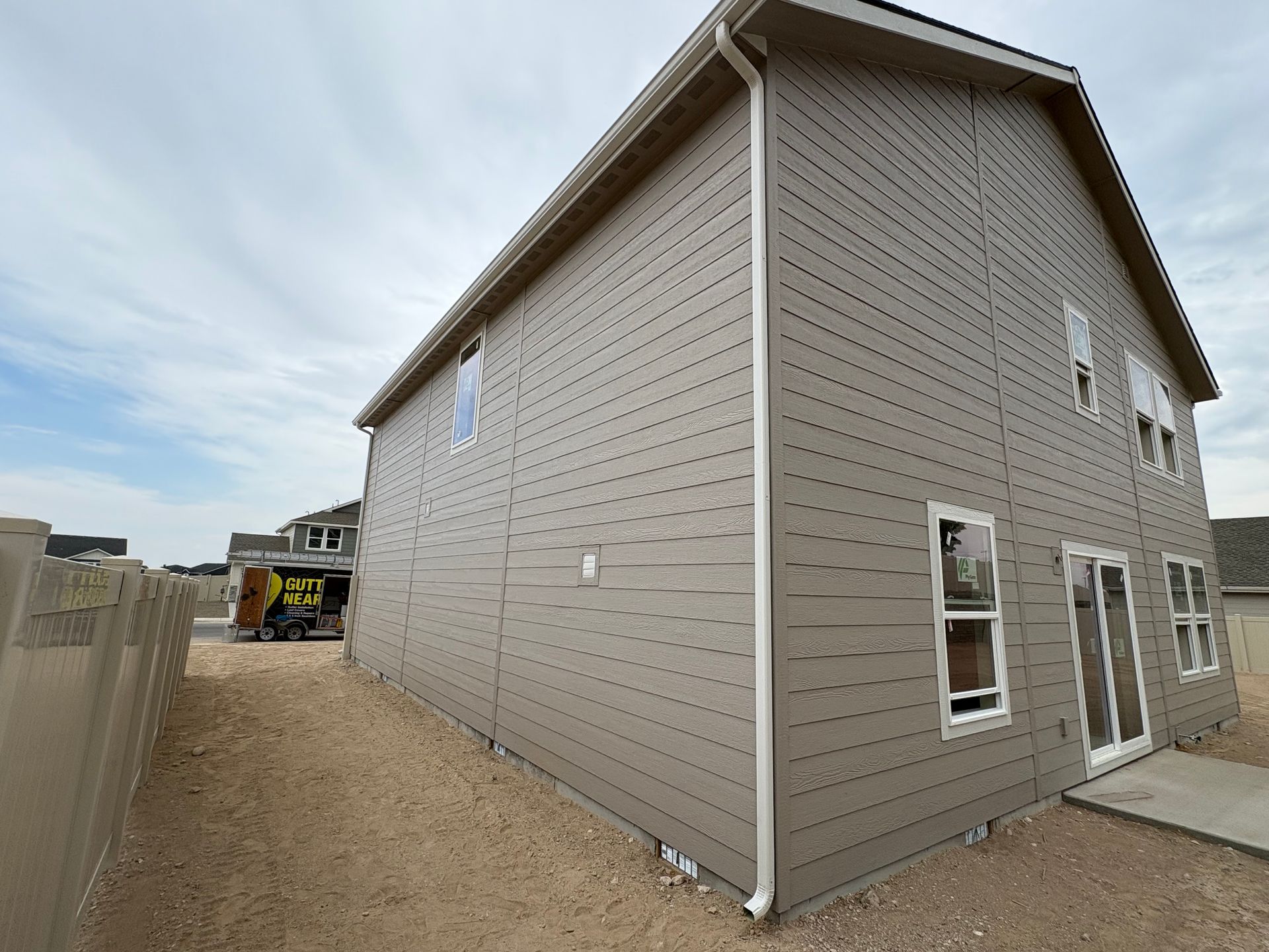 Two-story house with gray siding, white trim, and windows on a dirt lot.