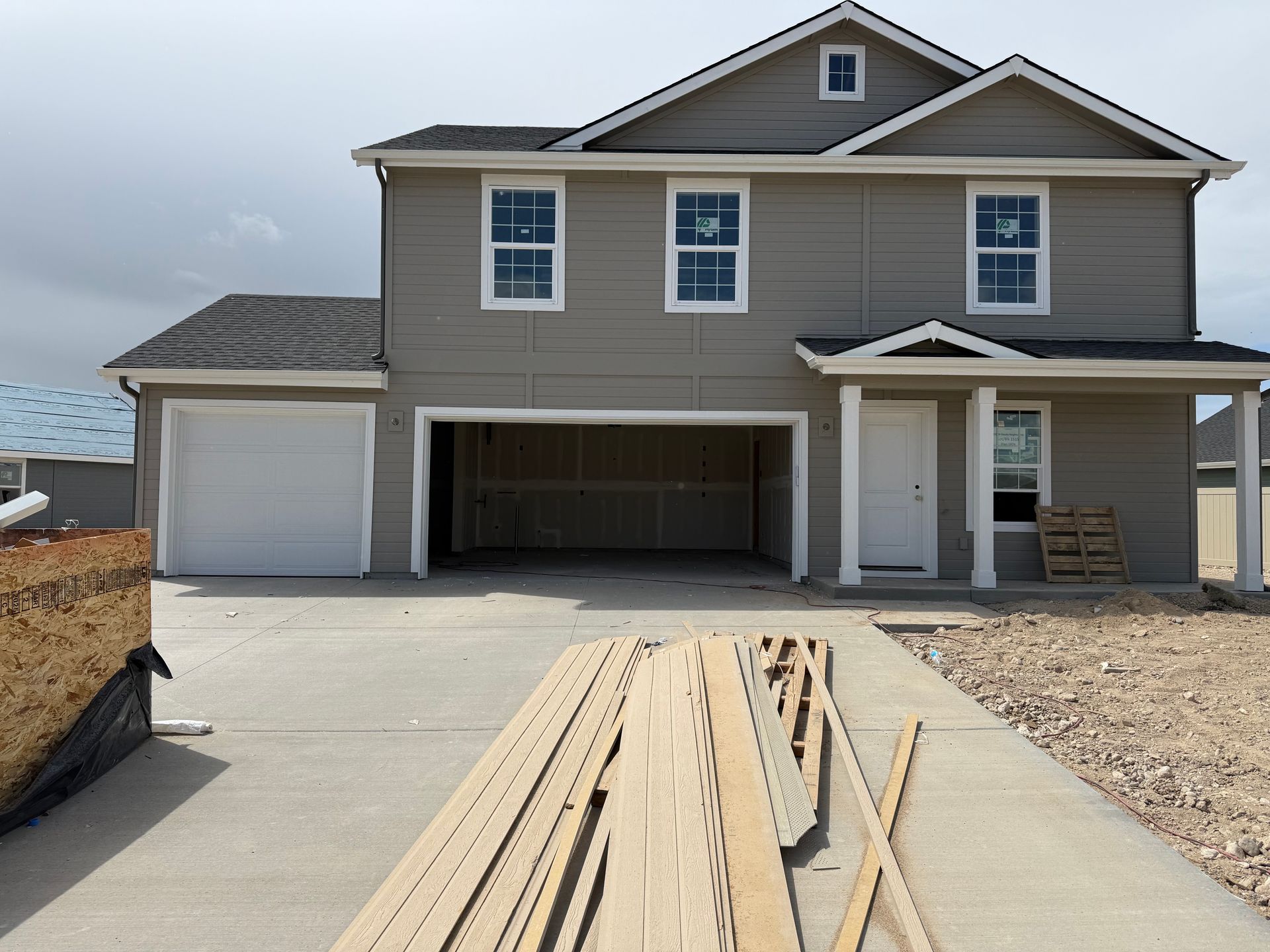 Two-story house with unfinished garage. Tan siding, white trim, gray roof, on concrete driveway with lumber.